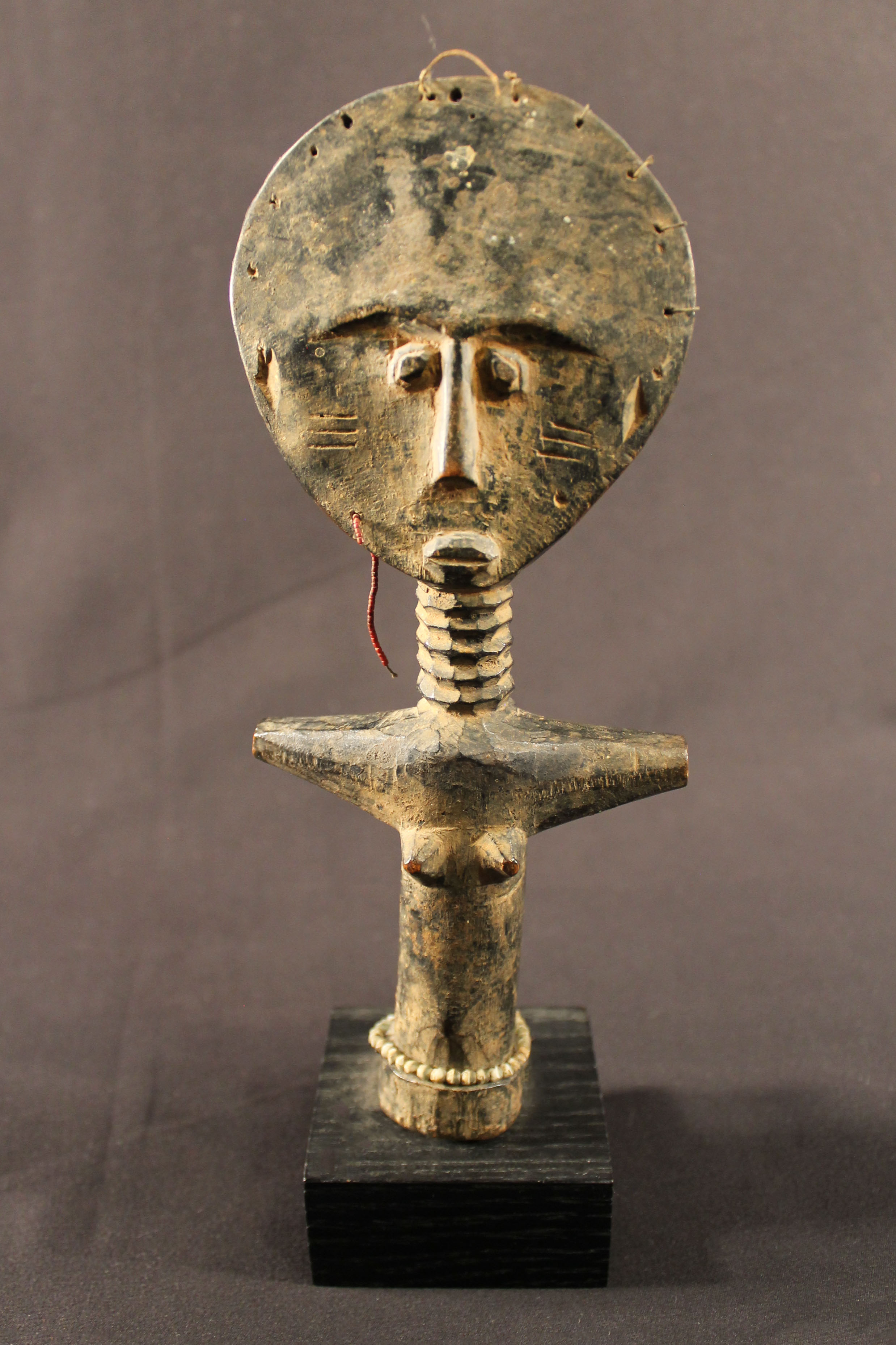 Carved wooden statue of a female figure in a cross pose with arms out. The woman has a large head with a string woven at the top. She has pointed breasts and is wearing waist beads. The statue sits on a dark wood base.