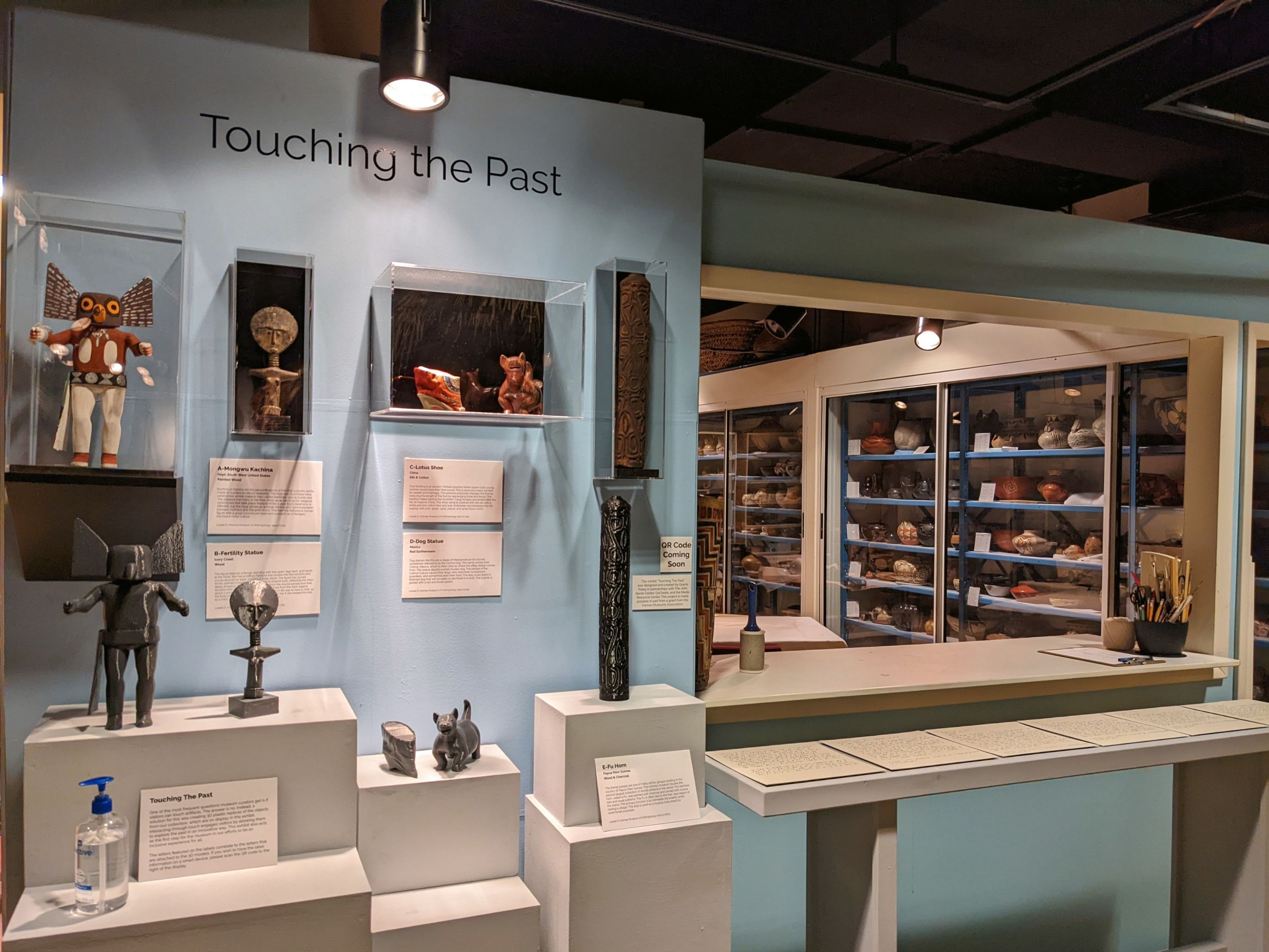The display has the 3D models sitting on a stand at arm level, with the actual artifacts mounted above them in clear cases attached to the wall. To the right of the exhibit is braille information on the objects, which are attached to a table that’s at arm level. The wall, the stands, and the table are all painted blue. 