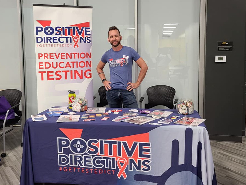 Person posing behind a table with information about Positive Directions and STI testing materials.