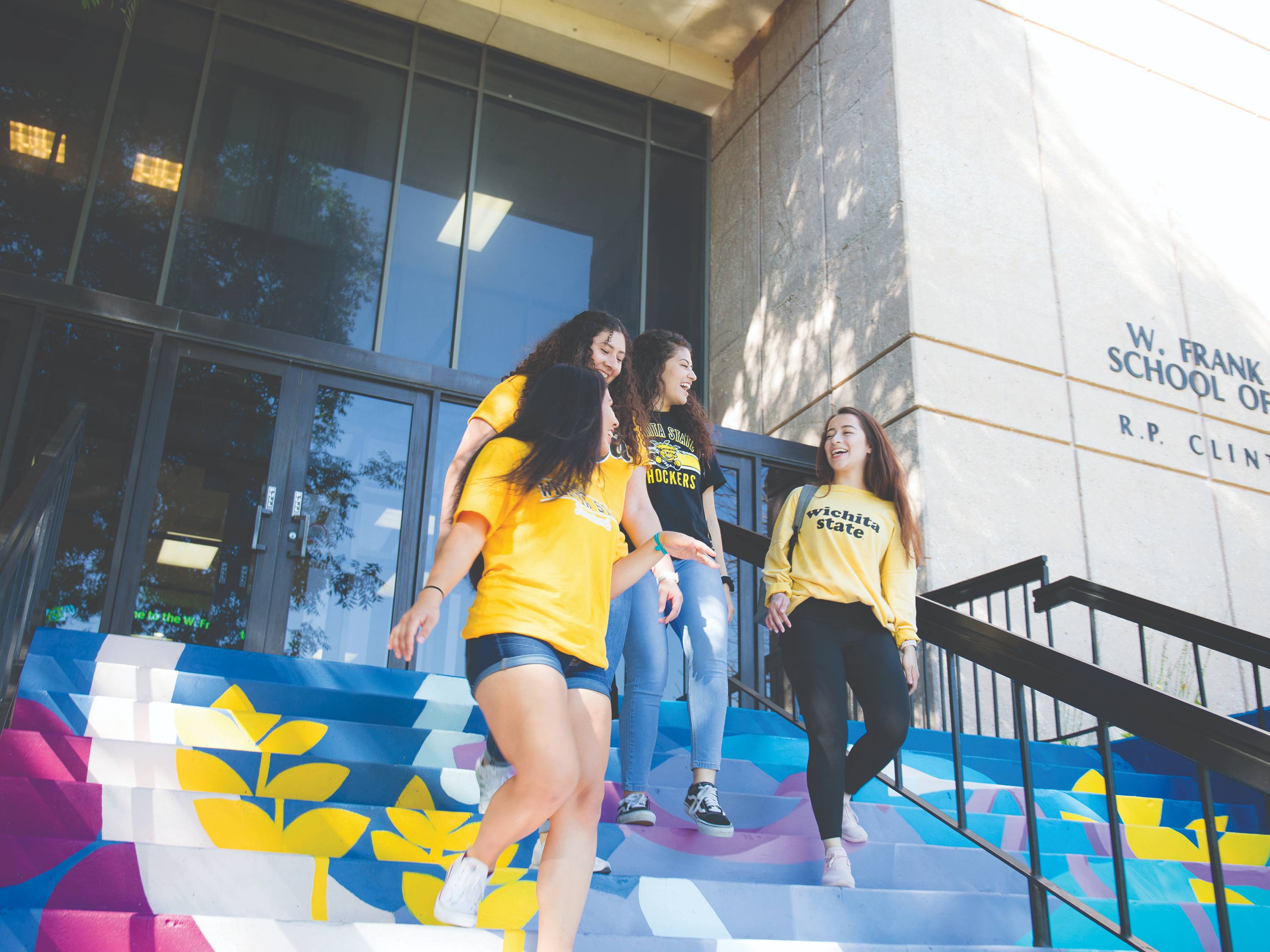 A group of female students descend the painted stairs at Clinton Hall, while laughing and chatting.