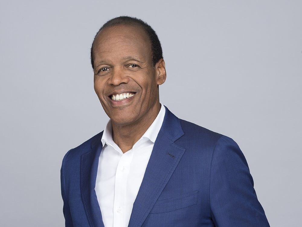 A man with a receding hairline and a bright smile, dressed in a blue blazer and white shirt, poses in front of a plain light gray backdrop.