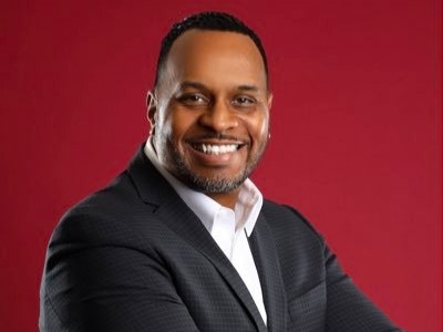 A man in a dark suit jacket and white shirt smiling against a red background.