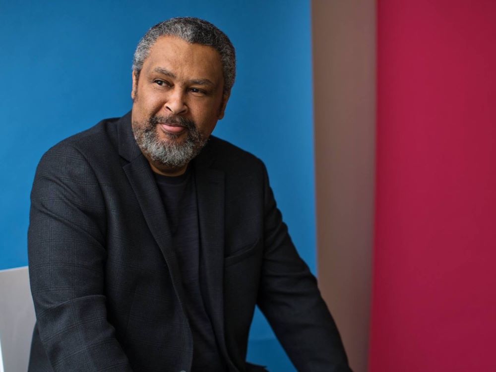 A man with gray hair and a beard, wearing a dark blazer over a black shirt, sits against a vibrant background divided into blue, beige, and red sections.