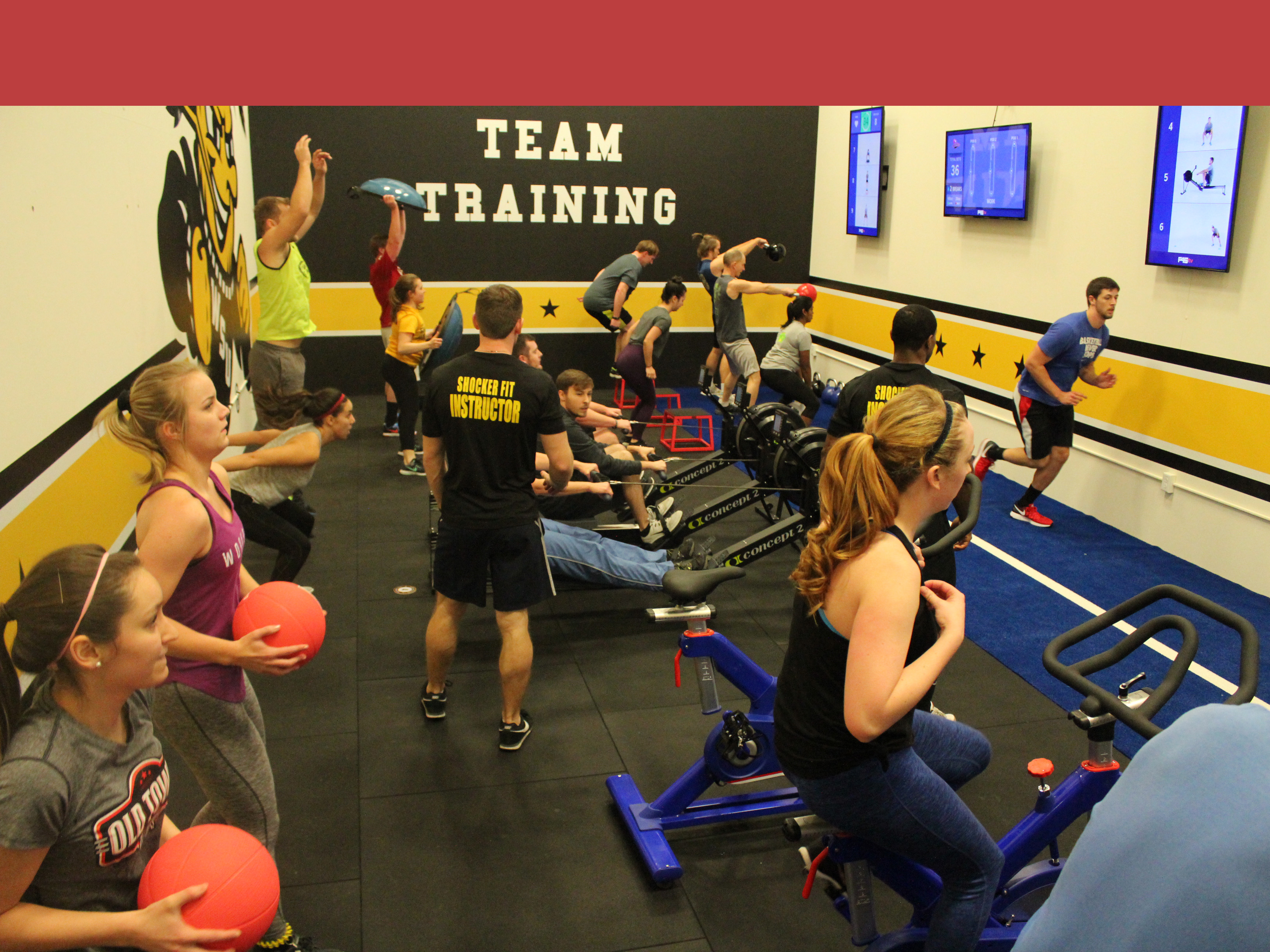 Group of people participating in F45 class. There are lots of excersizes going on, including bliking, medicine ball, kettleball, running, and rowing machine. the instructor is walking through this mess.