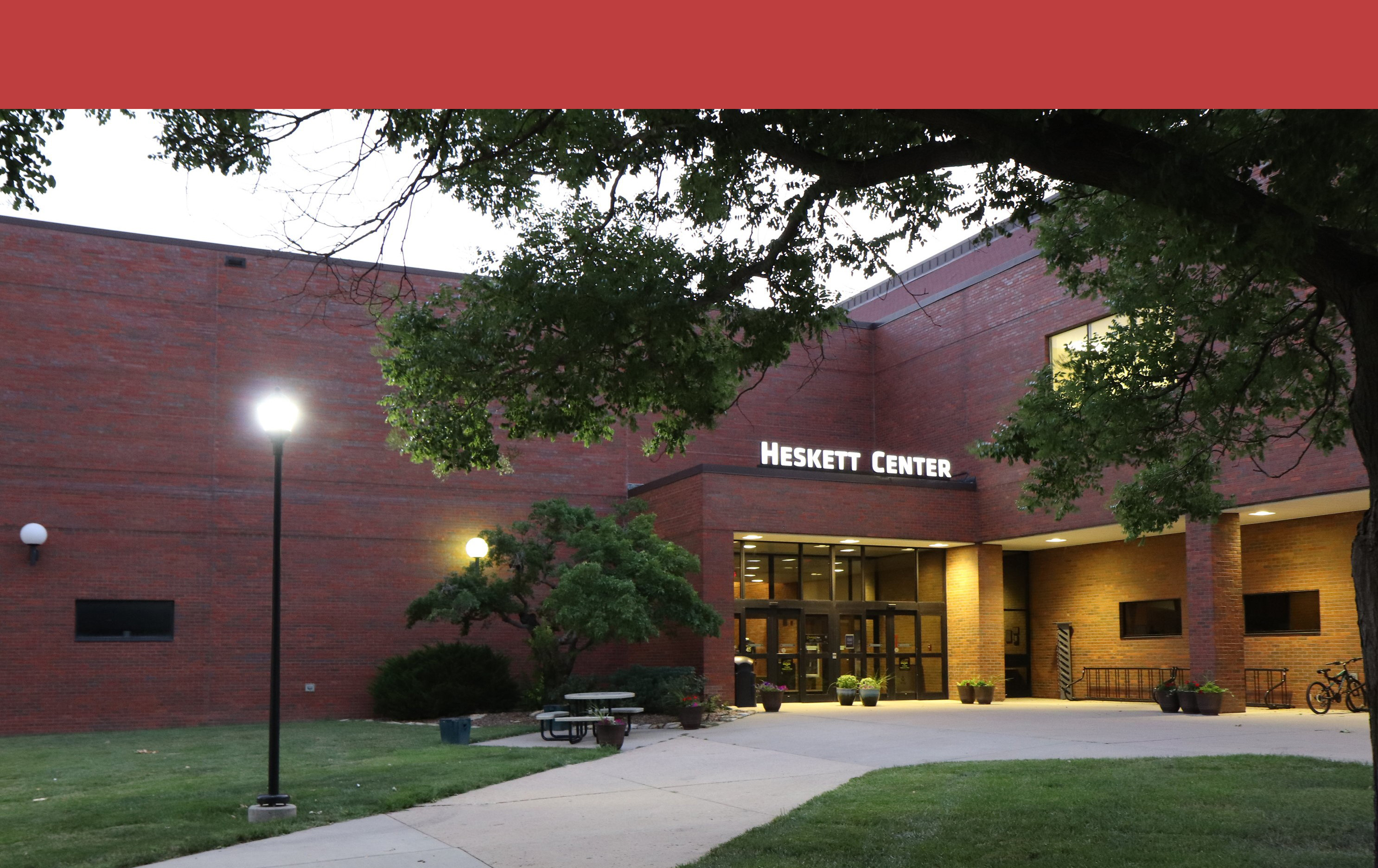 View of the Heskett Center from the outside. The sign above the door is lit up, as is the lamp post in the left of the image.
