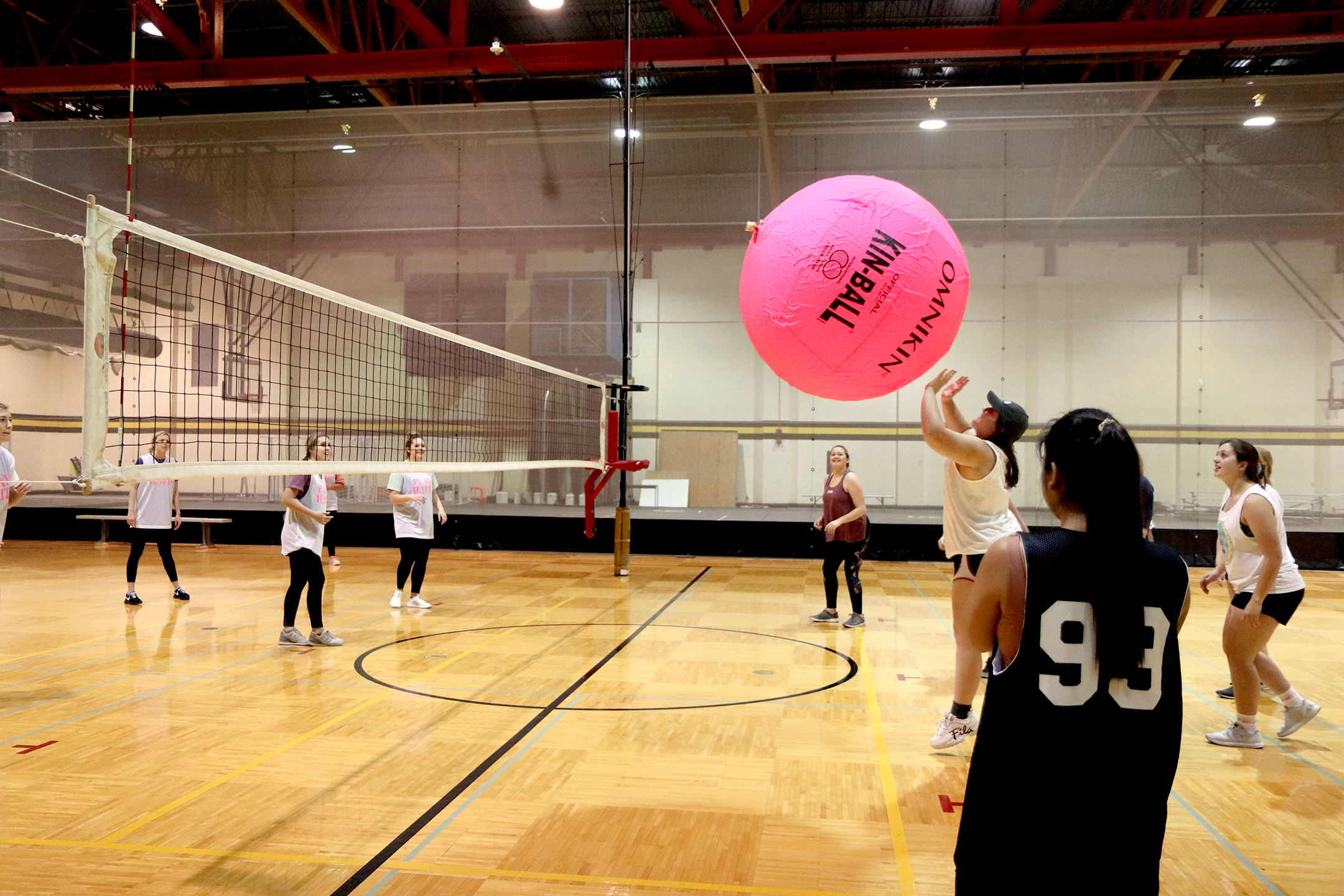 Group of people playing Big Pink Volleyball in the upstairs gym. Referee is on her feet, the ball is in the air between sides.