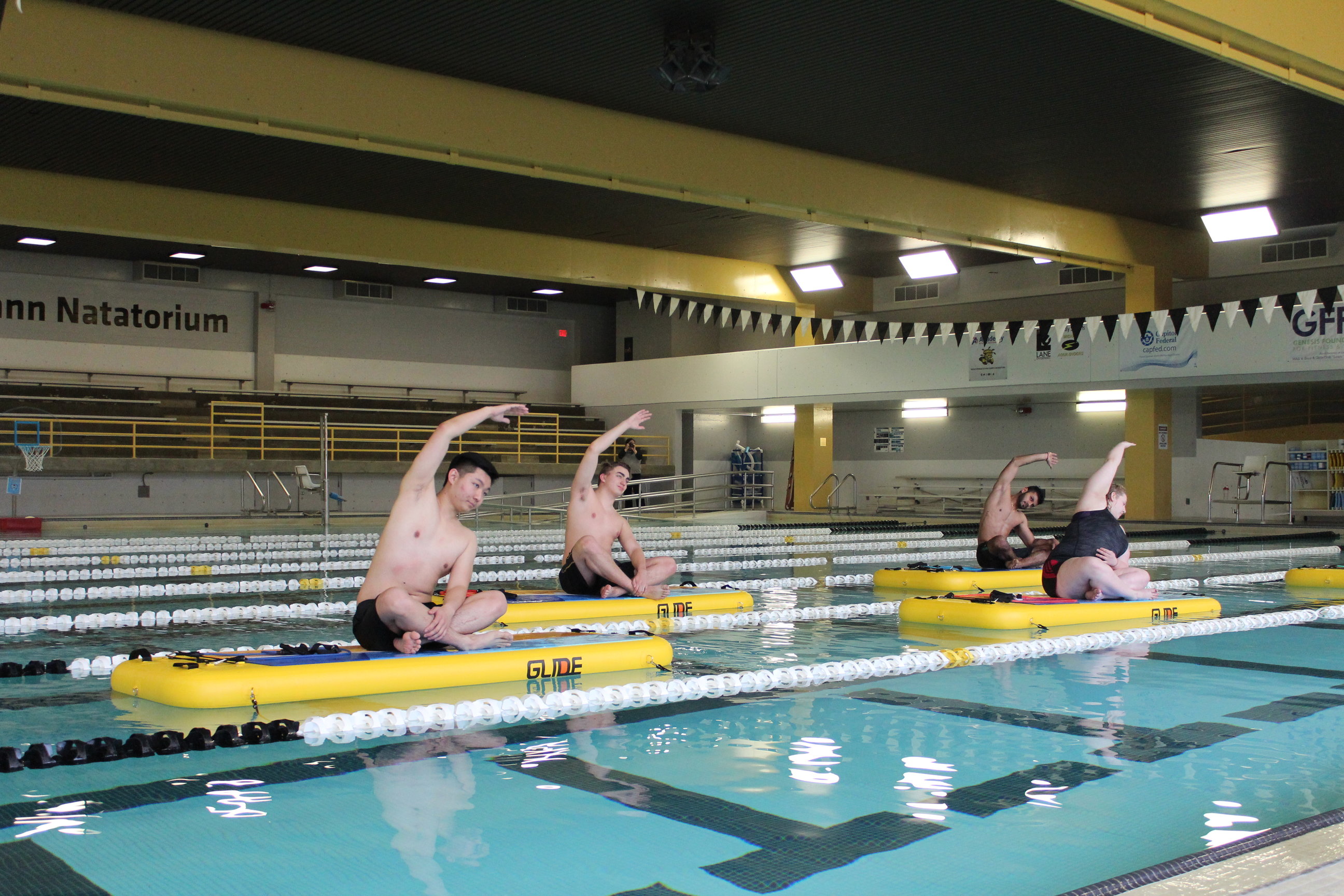 Four people sitting on GlideFit boards in the Wiedemann Natorium Pool. They are doing a yoga pose; sitting cross legged with one arm outstretched over their heads.