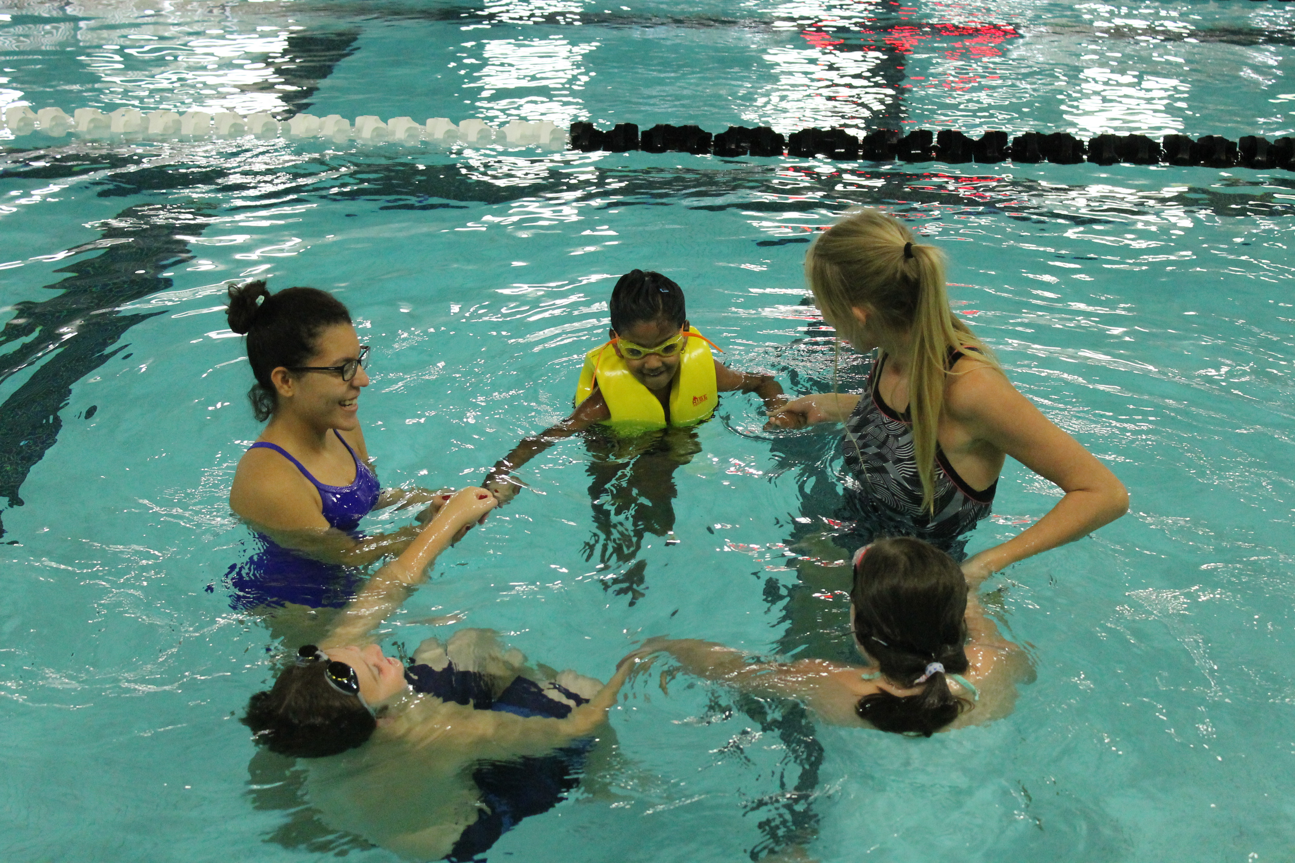 Two swim instructors hold hands with three instructees in a circular shape in the pool.