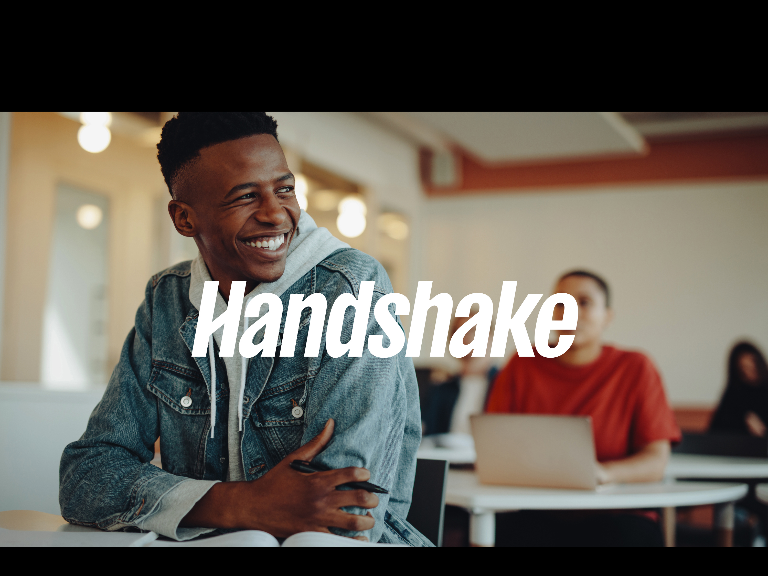 Image of student sitting in a classroom with an open notebook. Handshake logo overlaid on photo.