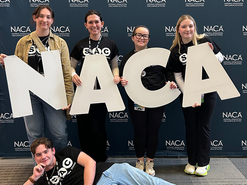 Five SAC students posing in front of a NACA backdrop and holding the letters N A C A.